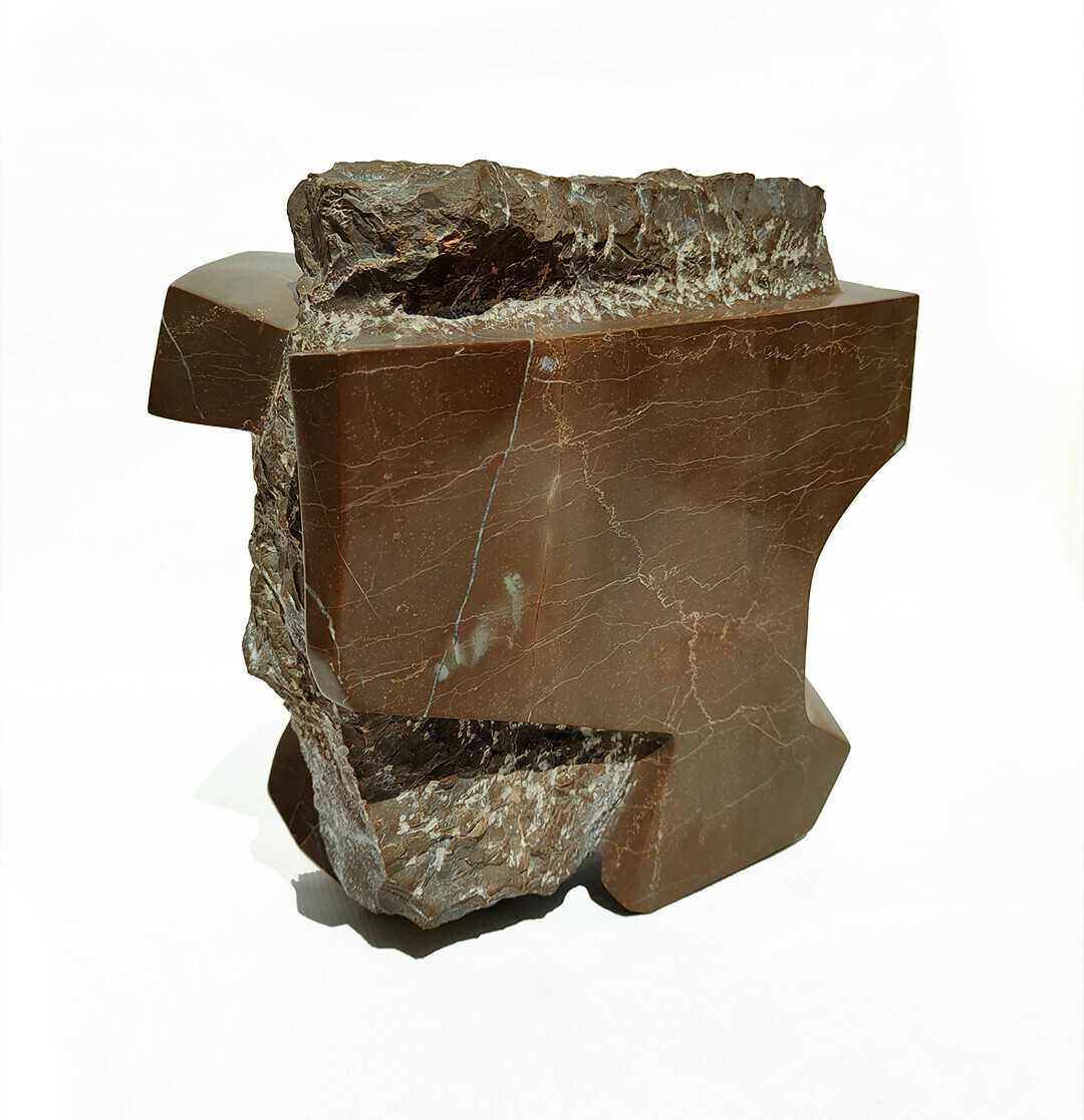 Front-right view of a stone carved sculpture made out of a single block of Grey Pulpis marble showing 2 visually distinct parts: 
- A central core, rough, amorphous, grey and shaped by hand with chisel and hammer, each strike aiming to provoke spontaneous breaks and accidental organic forms
- The core is partially covered by an external part made of geometrical shapes, planes and smooth curves, all of it modeled by a hand-held electrical grinder, sanded and waxed by hand, so its surfaces appear shiny and reflective dark brown with lots of clear veins and subtle changes in color 
- Both core and encasement, while being the same rock, are visually distinct and their forms and textures appear to react to each other