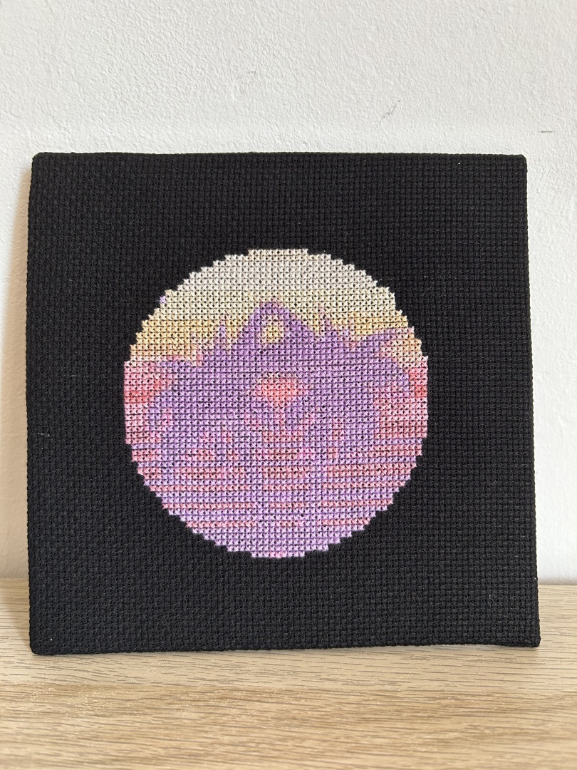 Square cross stitch on black Aida fabric of two palm trees with a sunset background in a circular design.