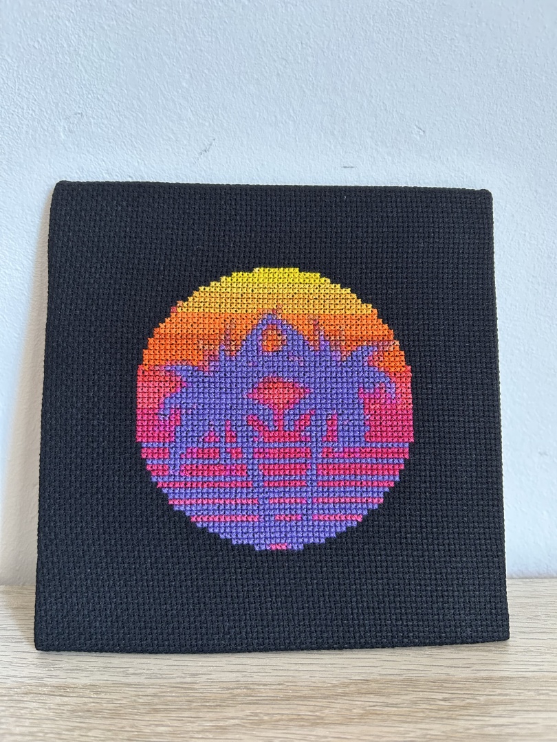 Square cross stitch on black Aida fabric of two palm trees in purple with a sunset background in shades of orange, pink and yellow in a circular design.