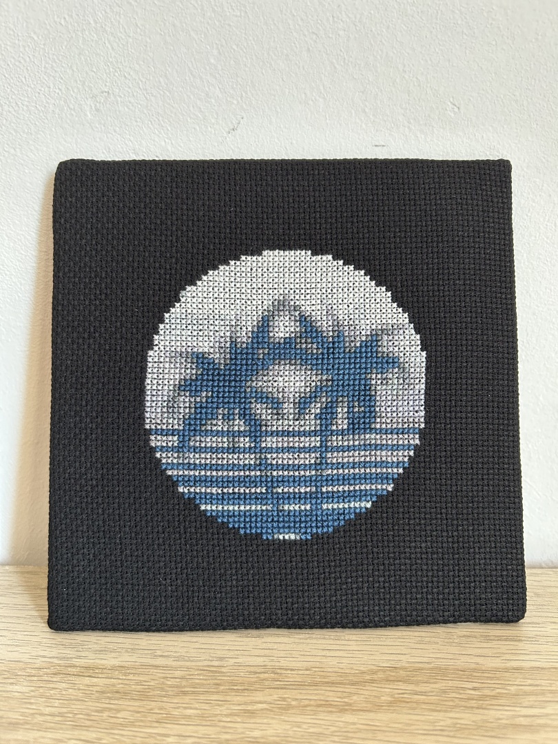 Square cross stitch of two palm trees with a sunset background in a circle on black Aida fabric. Design is in greys and cool blue hues giving a mono appearance.