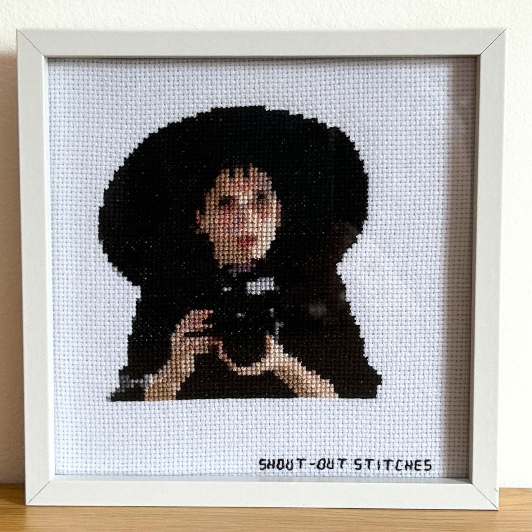 Lydia dressed in black with black sun hat holding her camera as she looks up at the window of the house where she see Adam and Barbara stitched on white aida fabric in a white square frame