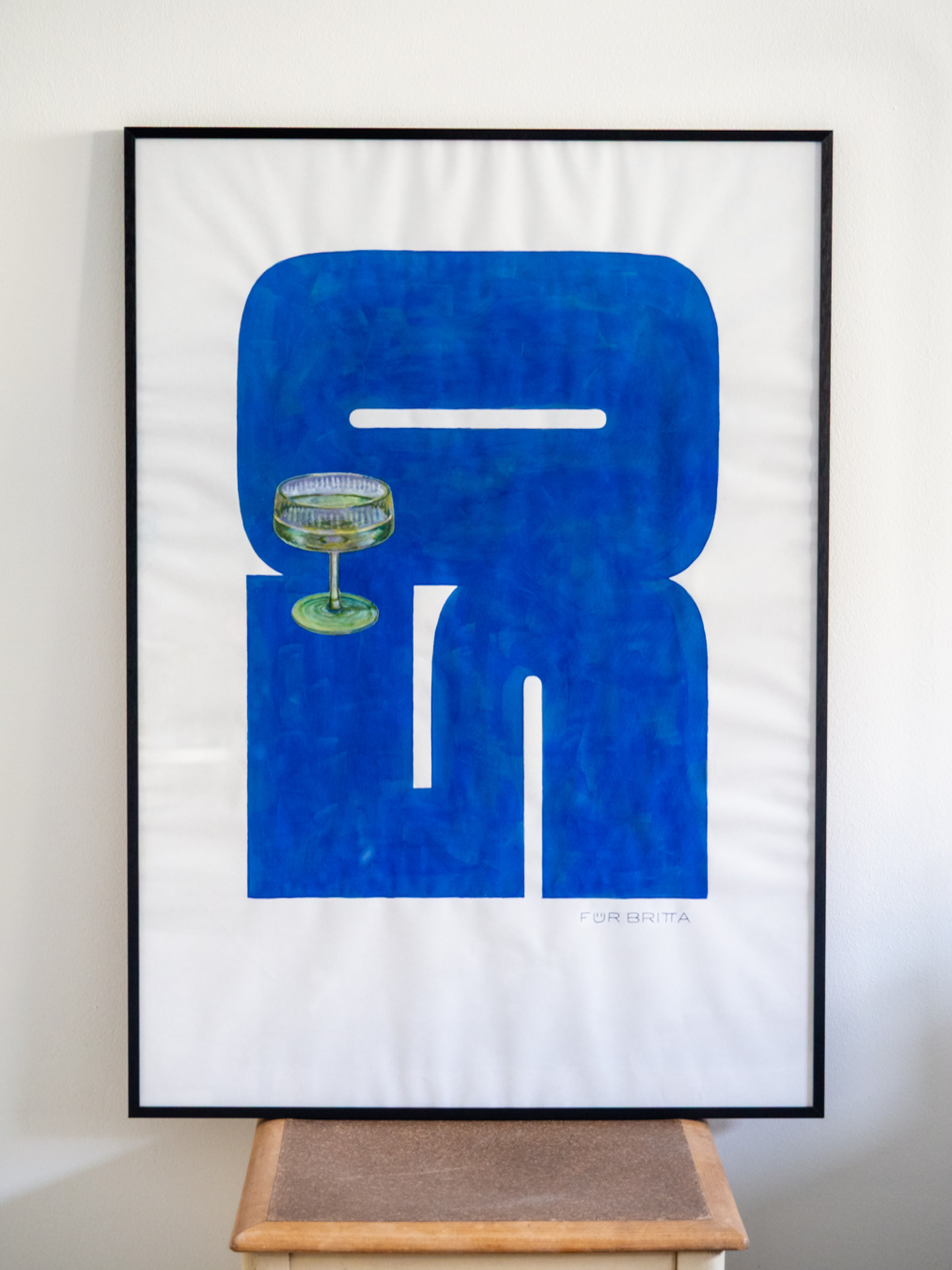 Hand painted poster in a frame depicting a large blue shape of the number 50 enclosing a glass of champagne.