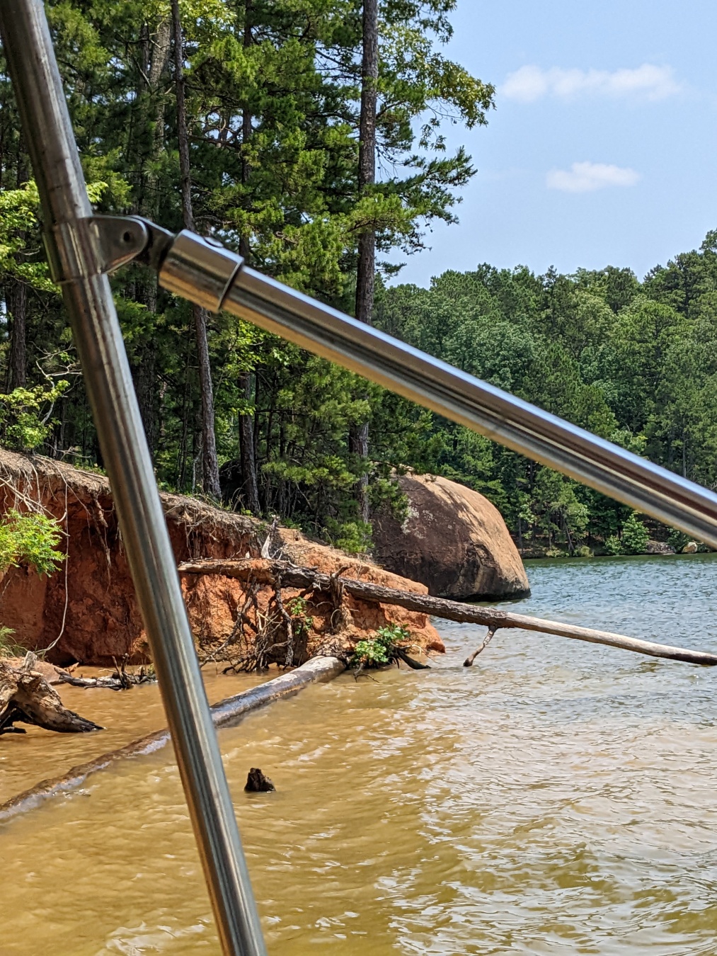 a photograph of a large rock in the water butting up against the land. the soil is orange and the trees are verdant. there's a metal cross brace in the foreground.
