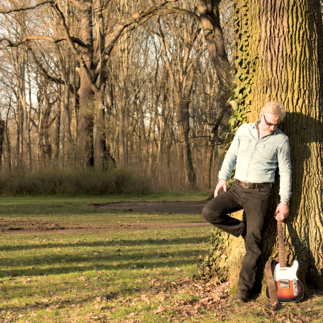 The main cover shot for the upcoming EP Simplicity by Andy Robson & The Urban Fox Network. Andy is leaning on a tree in a parc. He is looking down to his guitar leaning beside him. The sunlight gives the scene a soft and warm atmosphere.