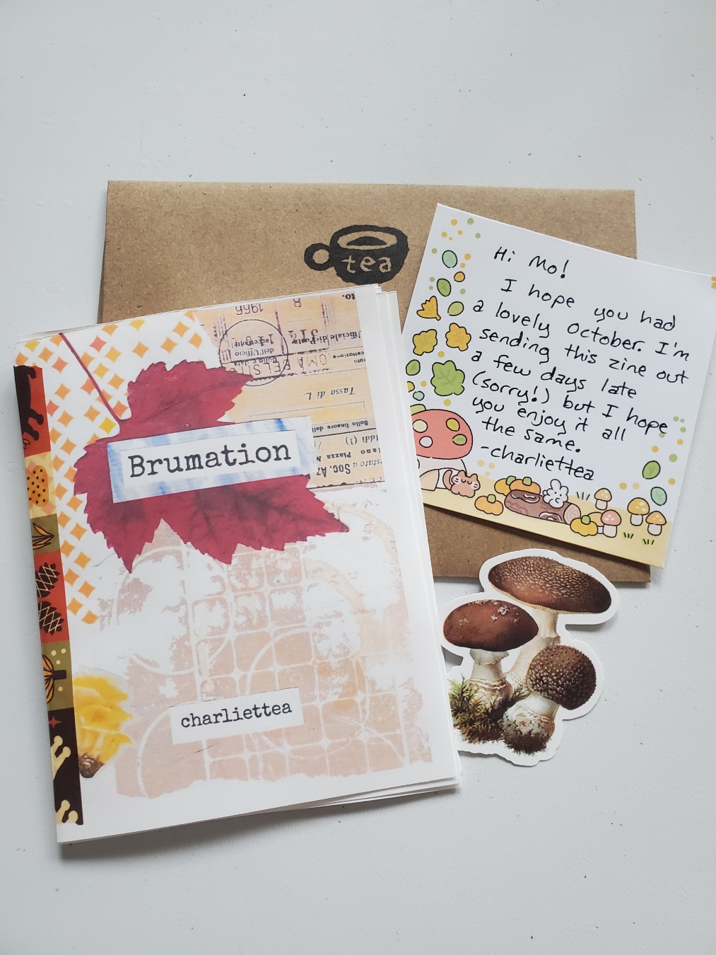 A photo of a zine, thank you note, envelope, and mushroom sticker. The zine is titled brumation and features a scan of a maple leaf on the cover. The envelope has a mug labeled tea stamped on it.