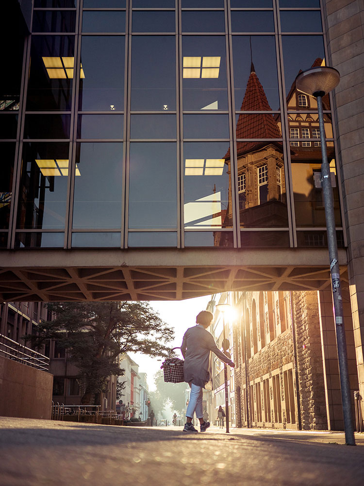 The bridge connecting the old and new town halls is lined with windows. Parts of the old town hall are reflected in them. A woman with a basket is walking under the bridge. The rising sun is reflected in a window that can be seen under the bridge on the right wall of the building. The picture has warm colors and is very rich in contrast.