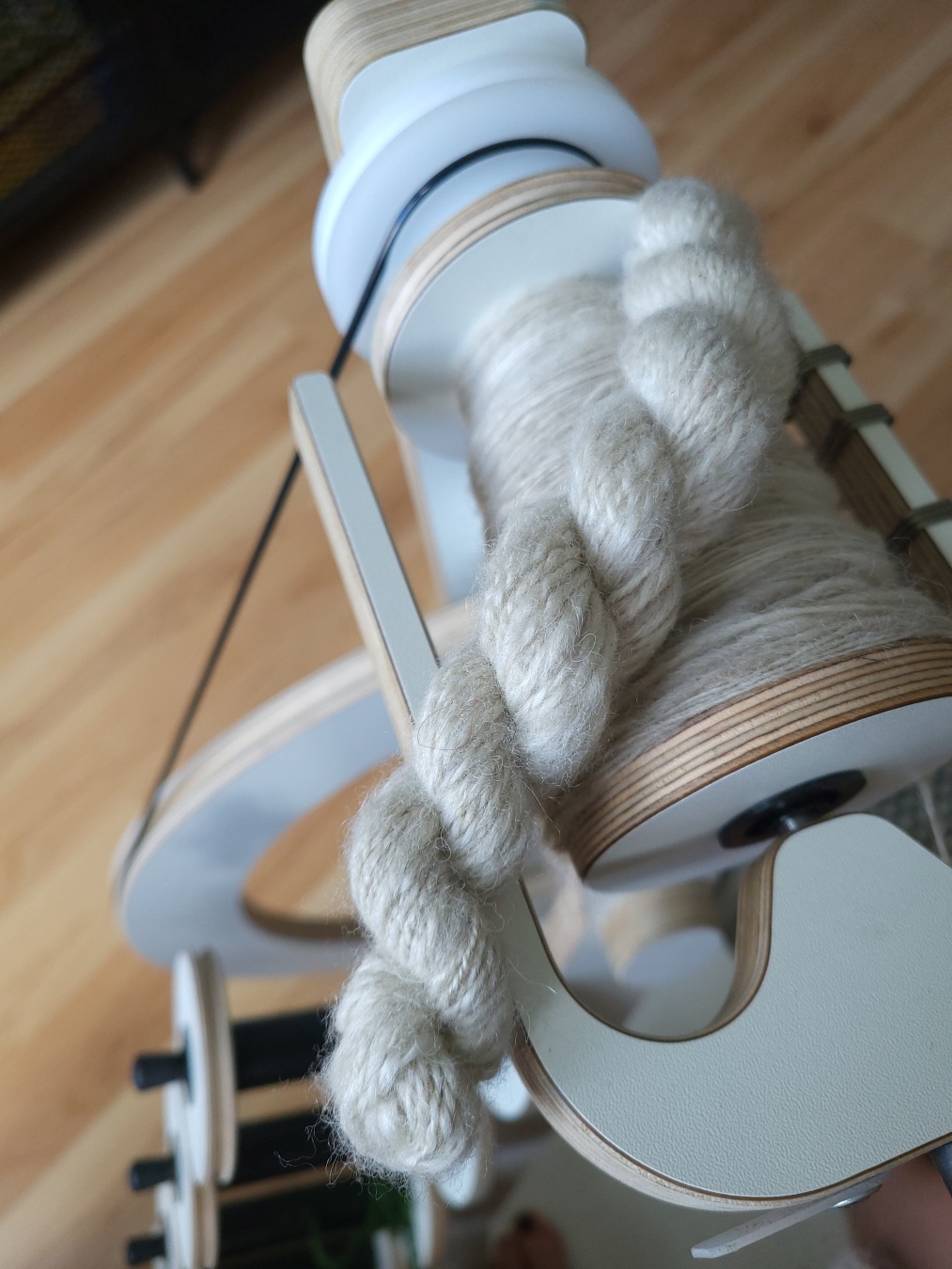 A modern white spinning wheel seen from above. On the bobbin and flyer lies a small white fluffy and drapey skein of yarn, the bobbin is filled with the same wool.