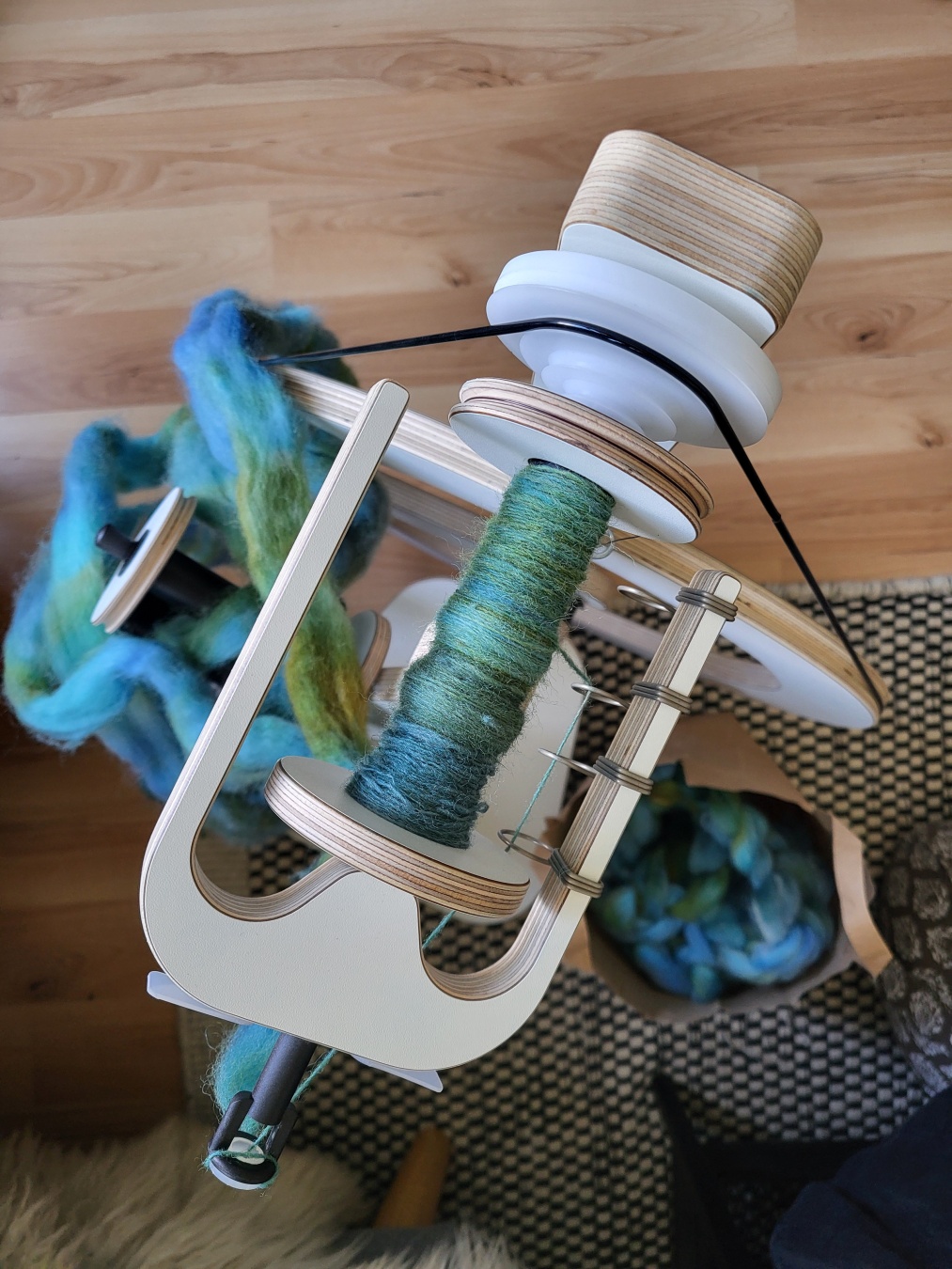 A white spinning wheel from above. You can see the bobbin in the flyer with a bit of blue-green yarn on it. Below, draped over the built-in lazy kate, is the source of the yarn: a predrafted commercial combed top in mostly blue and teal colours with some yellow, golden, green and brown parts and spots. Next to the wheel is a paper bag with even more of this top.