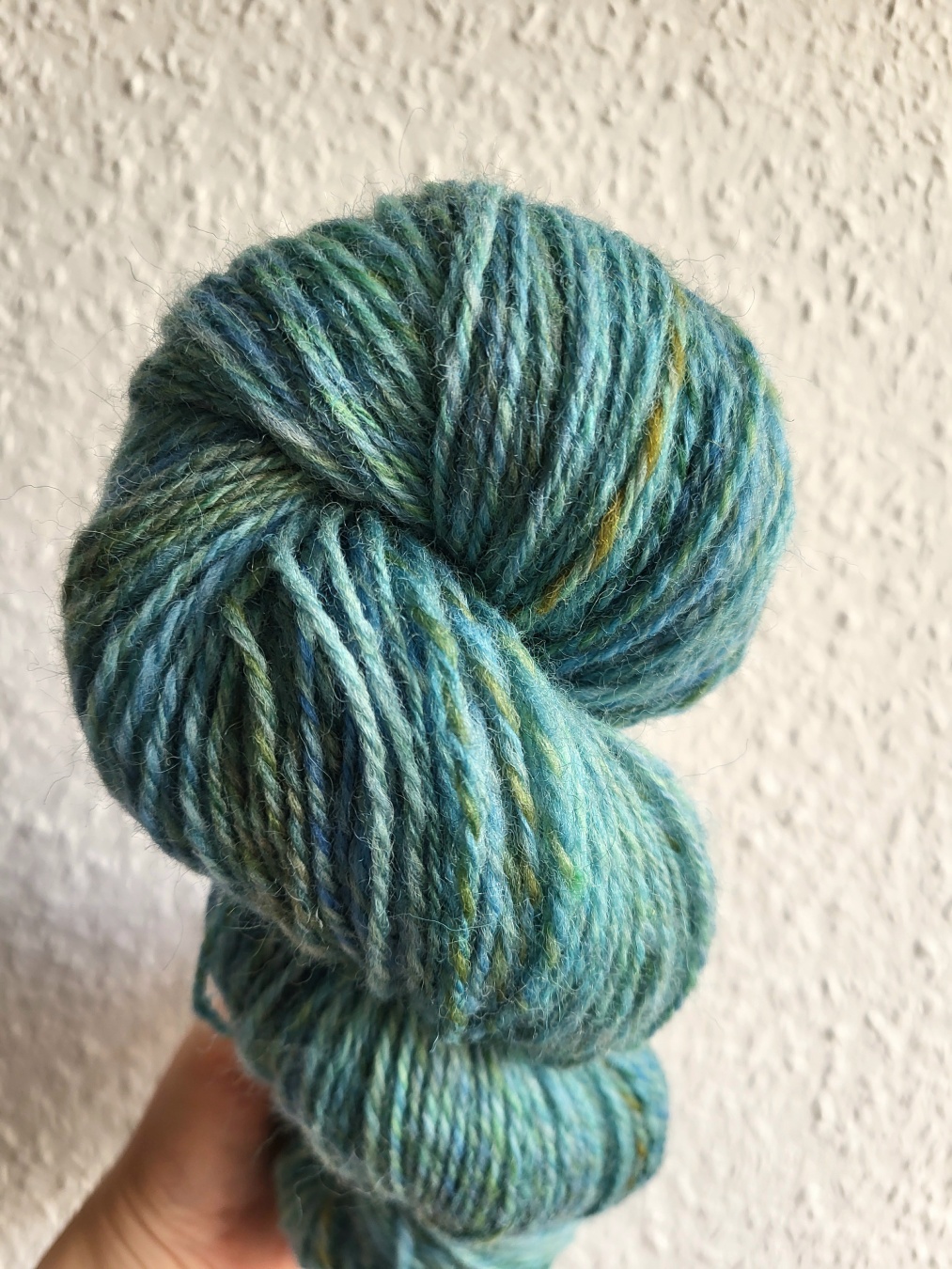 A skein of blueish yarn held by a hand in front of a white wall. The yarn is a 3-ply, so there is some colour variation in it with different blue and teal and green shades and some golden-green spots.