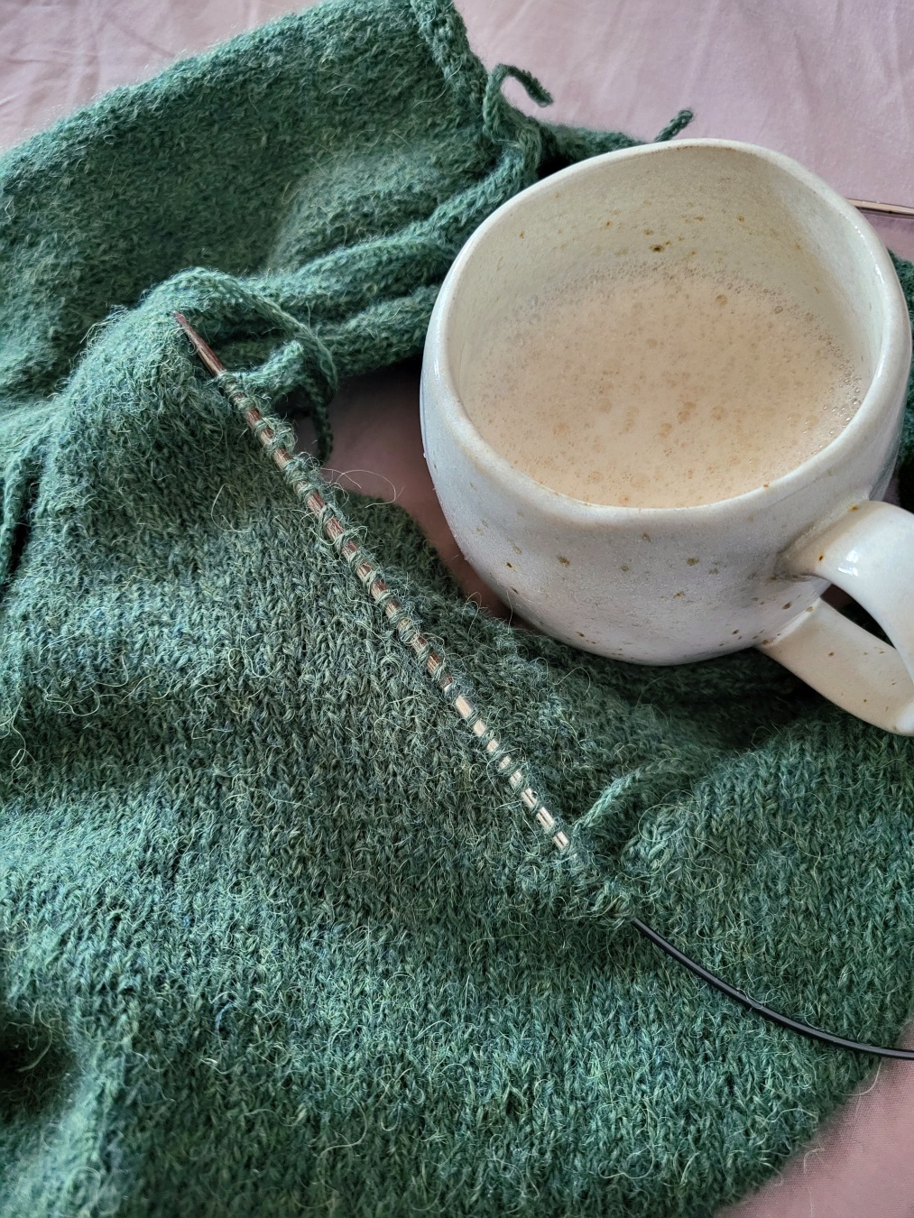 A dark green knit project in stockinette stitch. You cannot really see what it will become. A needle with picked up stitches sticks in the edge. Next to the project there is a rustic cup of cappuccino.