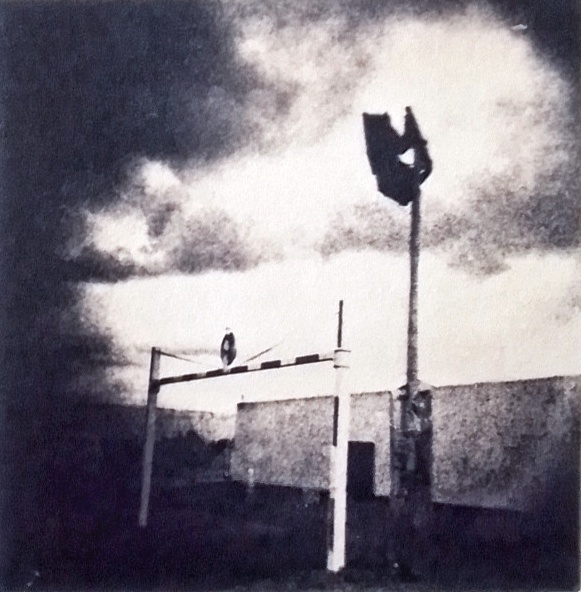 fotografía en alto contraste de un terreno en construction. se ben un à barrera y un poste de alumbrado. hay nubes gruesas en le cielo. la foto es un cyanotipo virado con café.