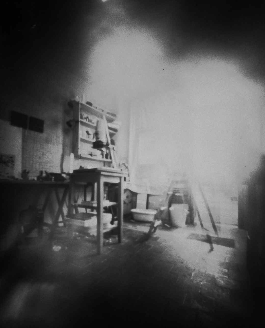 Fotografía estenopeica en blanco y negro. Se ve una mesa con una ampliadora. Atrás un estante y destellos de luz provenientes de la ventana.