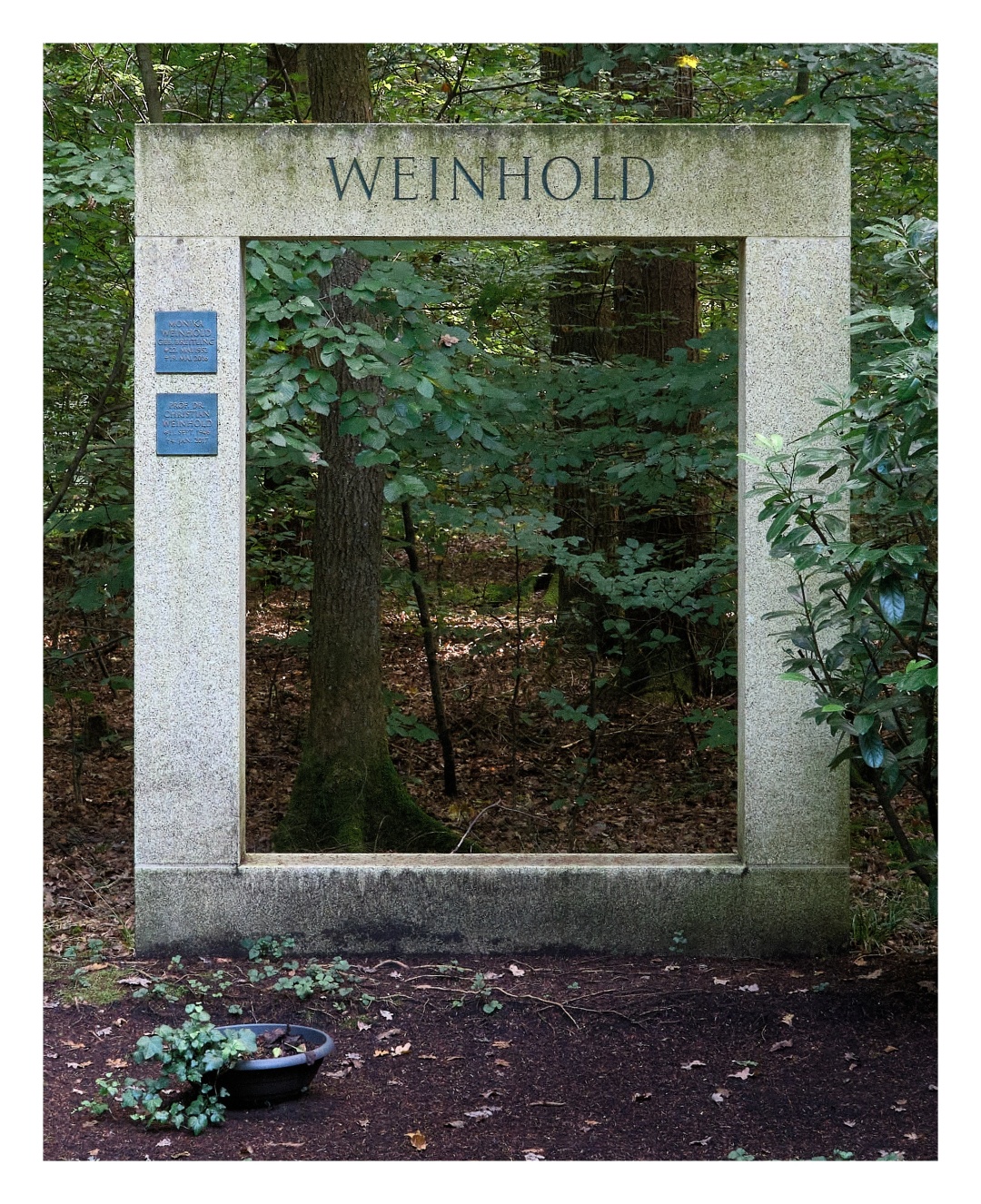 Das Foto zeigt einen Grabstein im Wald, der aus vier länglichen Steinquadern besteht. Der erste liegt quer auf der Erde, links und rechts auf ihm stehen zwei weitere Quader senkrecht. Oben liegt der vierte Quader quer. So ergibt sich ein Quadrat, das innen leer ist. Man kann wie durch einen Rahmen hindurchschauen und sieht Bäume, Blätter, Laub und Sonnenspiel.

The photo shows a gravestone in the forest consisting of four elongated stone blocks. The first lies horizontally on the ground, with two more blocks standing vertically on either side of it. The fourth block lies horizontally on top. This creates a square that is empty inside. You can look through it as if through a frame and see trees, leaves, foliage and the play of sunlight.
