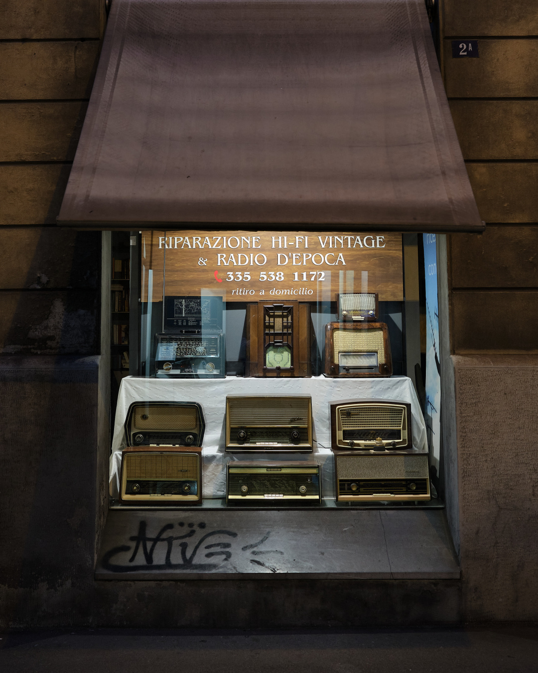Das Foto zeigt ein fast bodentiefes Fenster in einer italinienischen Hauswand bei Nacht. Eine nur fensterbreite Markise verdeckt den oberen Teil. Im Fenster ist Neonlicht. Ausgestellt sind auf drei Stufen neun sehr alte Radioapparate aus dem letzten Jahrhundert. Auf einem selbstgebastelten Schild darüber steht: "RIPARAZIONE HI-FI VINTAGE & RADIO D'EPOCA"

The photo shows an almost floor-to-ceiling window in the wall of an Italian house at night. An awning covering only the width of the window conceals the upper part. There is neon light in the window. Nine very old radio sets from the last century are displayed on three steps. A homemade sign above them reads: ‘RIPARAZIONE HI-FI VINTAGE & RADIO D'EPOCA’