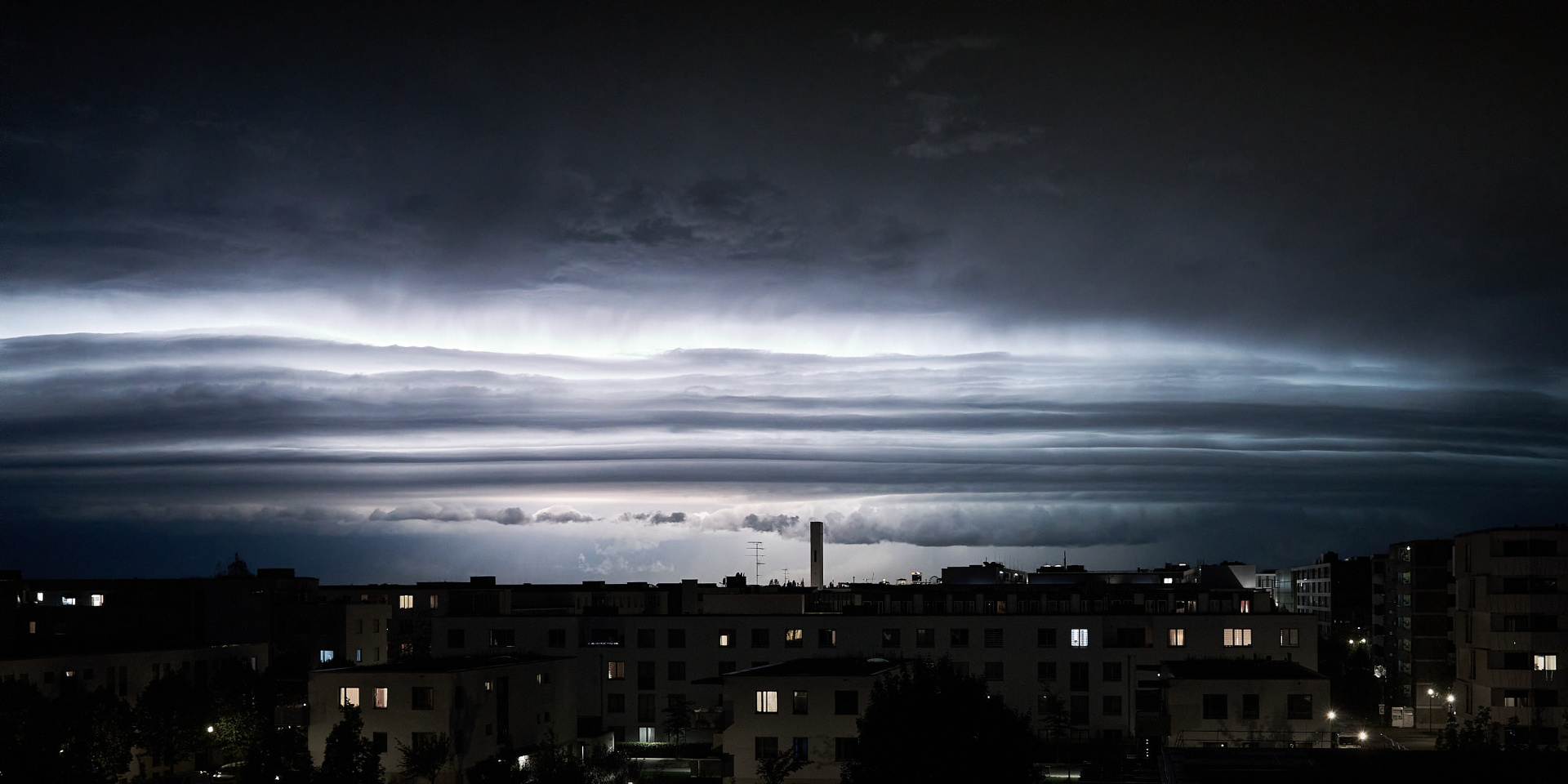 Foto bei Nacht von oben auf eine dunkle städtische Wohngegend unter dem Nachthimmel. Das Foto teilt sich horizontal in drei Streifen: unten (erstes Drittel) vierstöckige Häuser mit Flachdach, ein paar Fenster sind erleuchtet, manche mit warmem Licht, manche mit kaltem, ein par Bäume zwischen den Häusern. Darüber ein blauschwarzer Nachthimmel. Im mittleren Drittel zerreißt ein gleißendes Lichtband quer über das ganze Foto den Himmel. Das nächtliche Wetterleuchten zeigt ein heranziehendes Gewitter an.

Nighttime photo taken from above of a dark urban residential area under the night sky. The photo is divided horizontally into three strips: at the bottom (first third) are four-story houses with flat roofs, a few windows are lit, some with warm light, some with cold light. Above is a blue-black night sky. In the middle third, a glistening band of light across the entire photo tears through the sky. The nocturnal lightning flashes indicate an approaching thunderstorm.